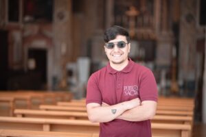 Foto di un ragazzo che sorride con le braccia incrociate all'interno di una Chiesa. Guarda verso la fotocamera.