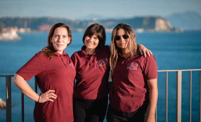 tre ragazze, abbracciate tra loro, guardano verso la fotocamera sorridendo. dietro di loro una ringhiera che affaccia sul golfo di Napoli