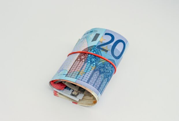 rotolo di soldi con attorno un elastico rosso, banconota più all'esterno di venti euro, sfondo bianco