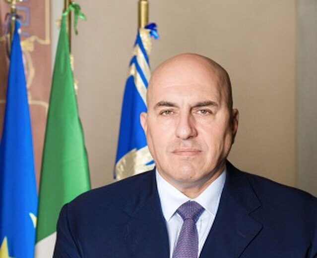 Guido Crosetto guarda la fotocamera. dietro di lui tre bandiere, tra cui presumibilmente quella italiana e quella dell'unione europea. è vestito elegantemente, con giacca blu, cravatta viola e camicia bianca. sguardo serio, stanza illuminata.